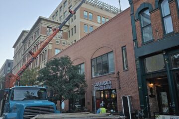 Bill Boom crane truck with its boom extended over a two-story brick building on Blake Street in downtown Denver, lifting a rooftop HVAC unit
